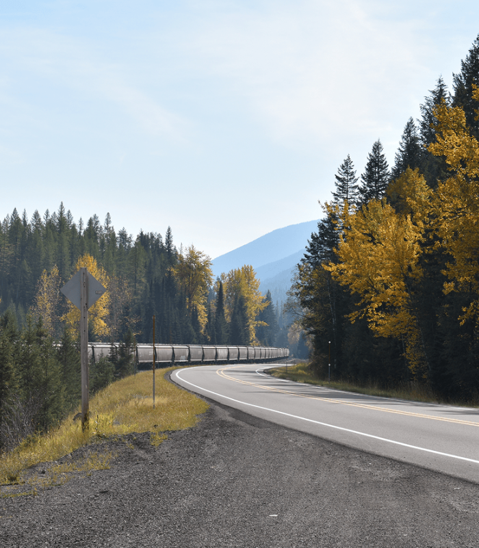 Empty road surrounded by green and yellow fall trees and alongside a train in the distance.
